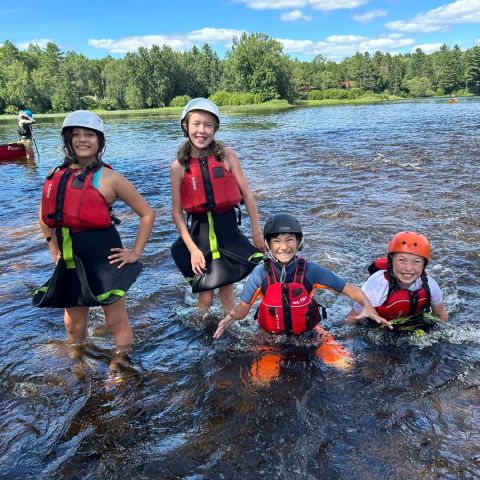 Four kids wearing helmets and life jackets stand in a shallow river, smiling under a sunny sky.