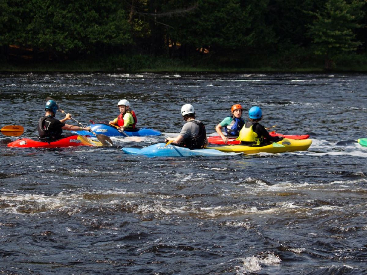 Five people kayaking on a river wearing helmets and life jackets, with trees in the background.
