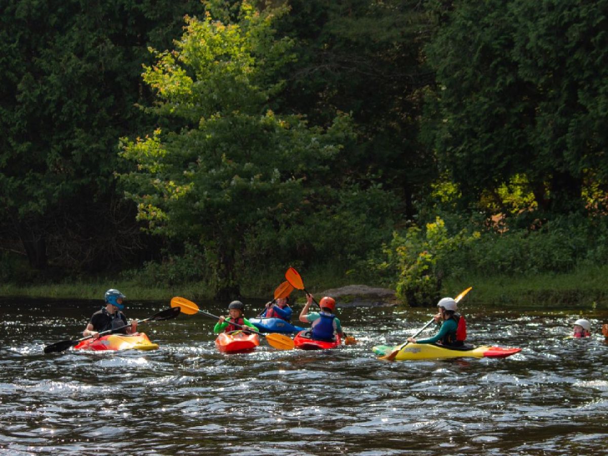 Group of kayakers with helmets paddling on a river surrounded by trees.