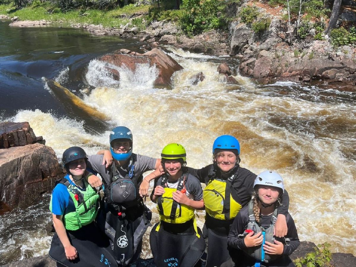 Group wearing helmets and life jackets posing by a river with rapids.