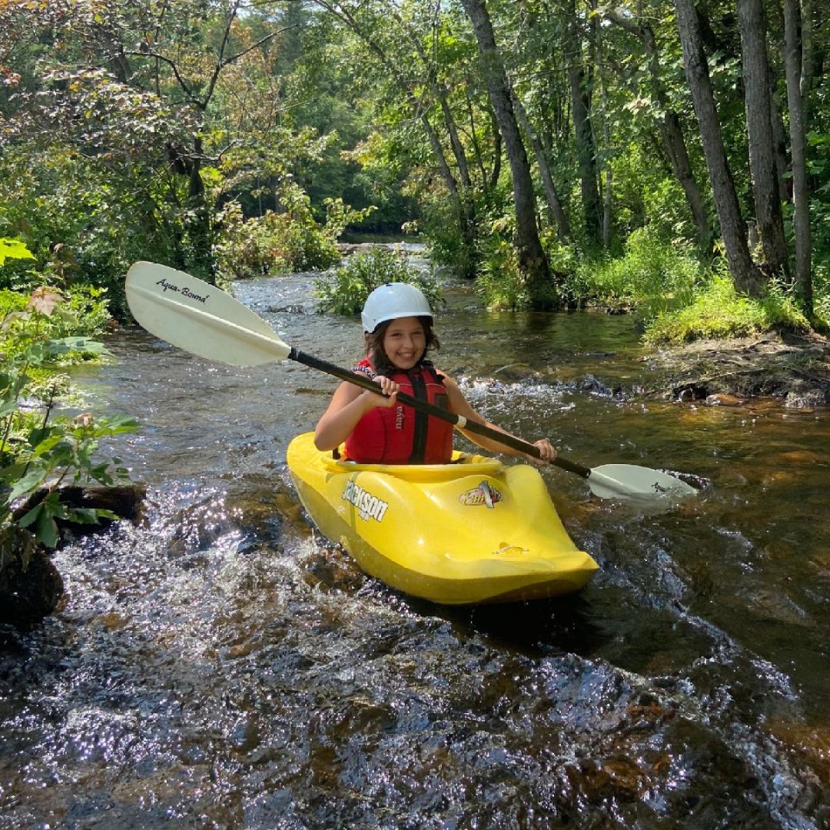 Person kayaking on a small stream in a forested area with green foliage.