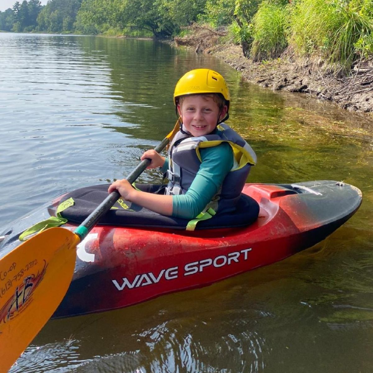 Child kayaking on a river, wearing a yellow helmet and life vest, with trees in the background.