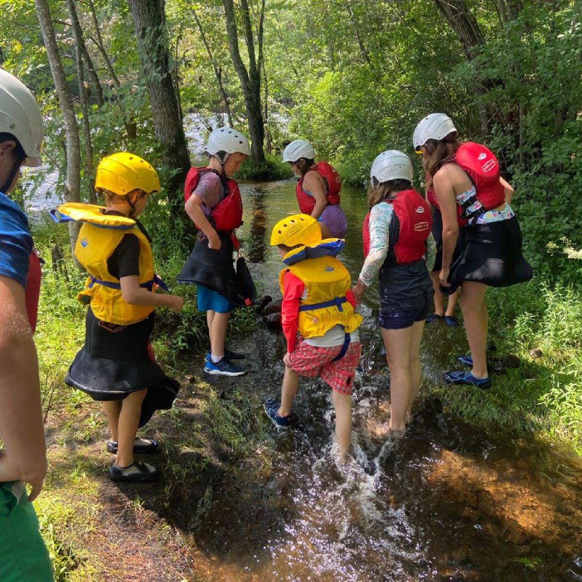 Children wearing helmets and life vests wade through a shallow stream in a wooded area.
