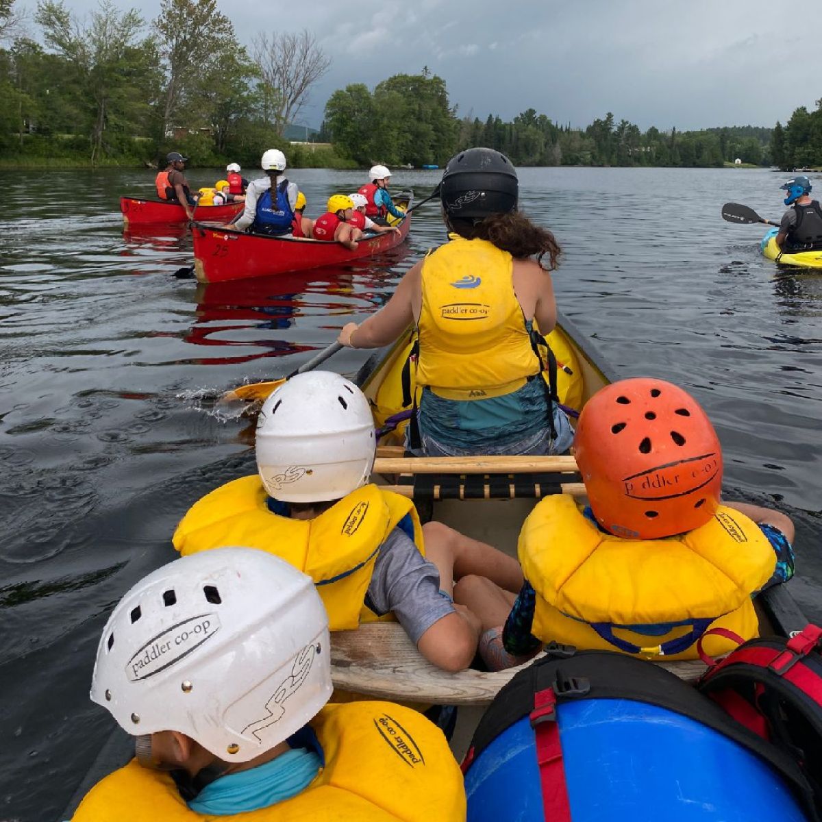 People in helmets and life vests paddling canoes on a lake with trees in the background.