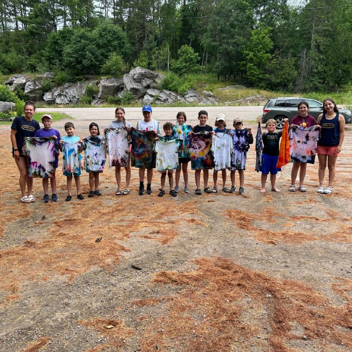 Group of kids holding colorful tie-dye shirts outdoors with trees in background.