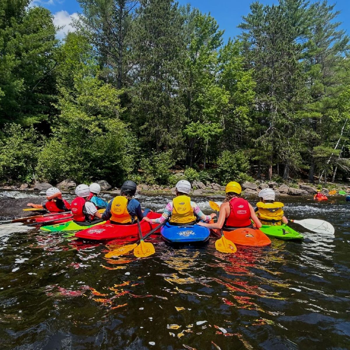 Six people in colorful kayaks on a river, surrounded by green forest, wearing helmets and life vests.