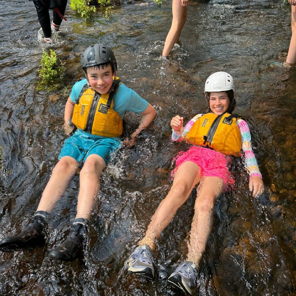Two kids wearing helmets and life jackets sit in a shallow creek, smiling.