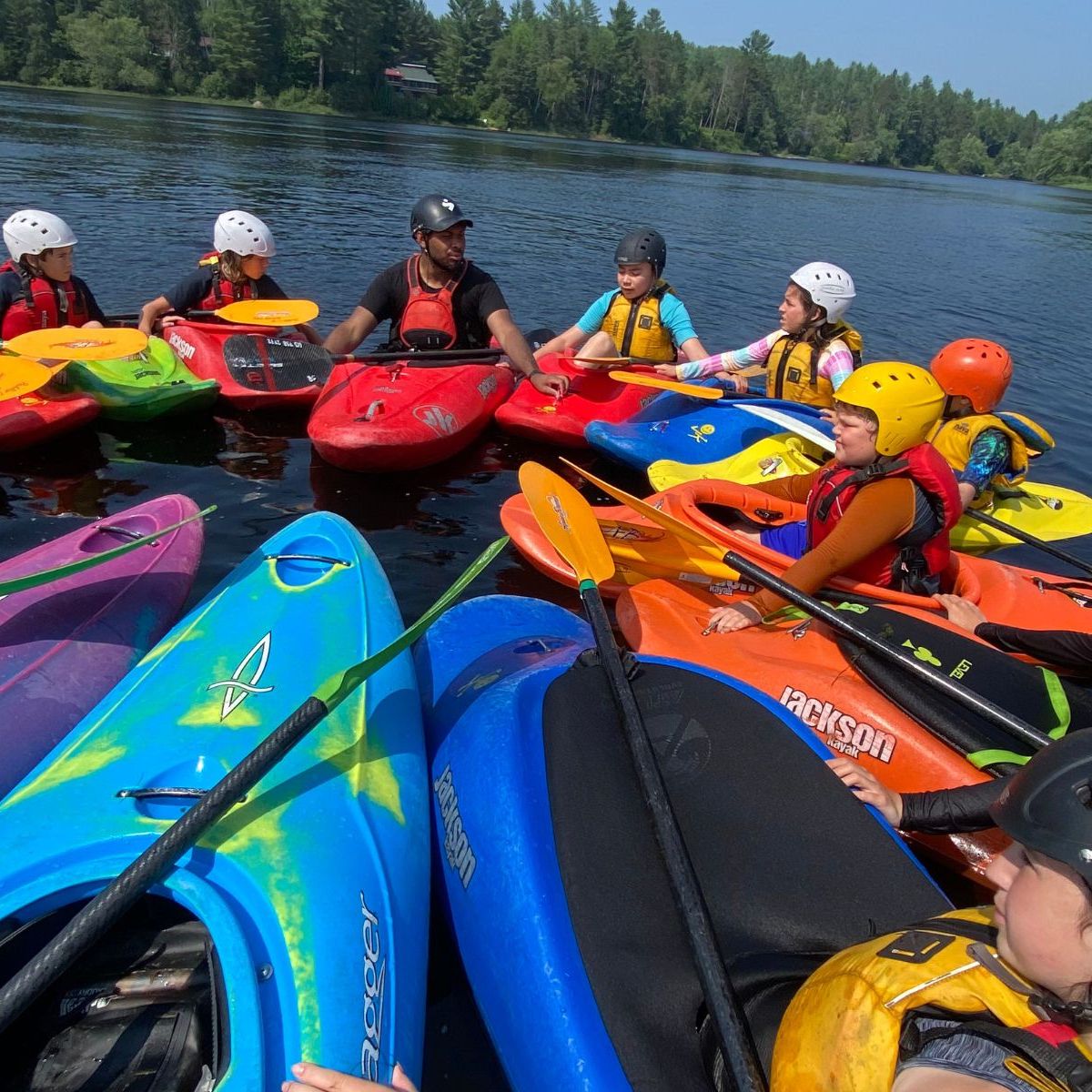 Group of kayakers in colorful kayaks gathered on a lake, wearing helmets and life vests.