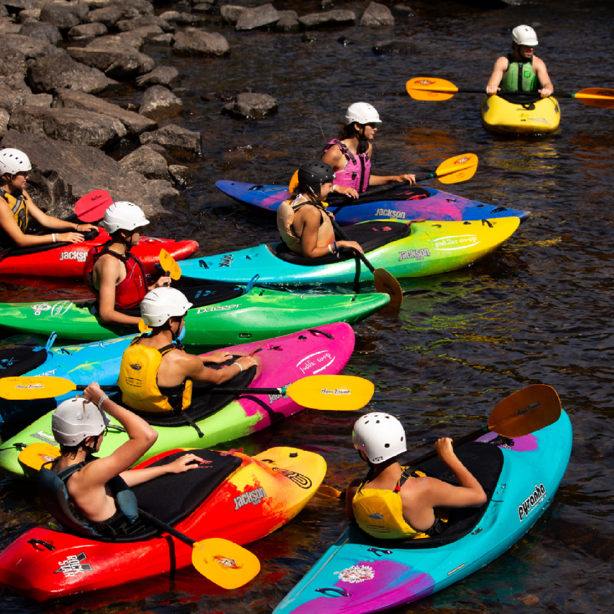 Group of people kayaking in colorful boats on a river wearing helmets and life vests.