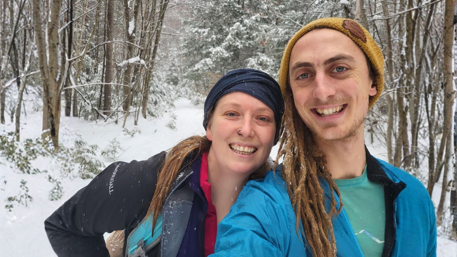 Two people smiling in a snowy forest wearing winter jackets and head coverings.