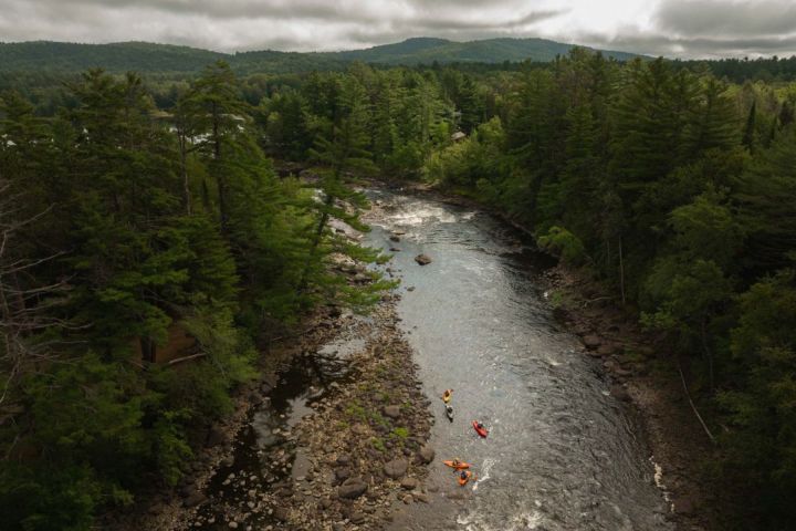 Three kayakers on a river surrounded by dense forest under cloudy skies.