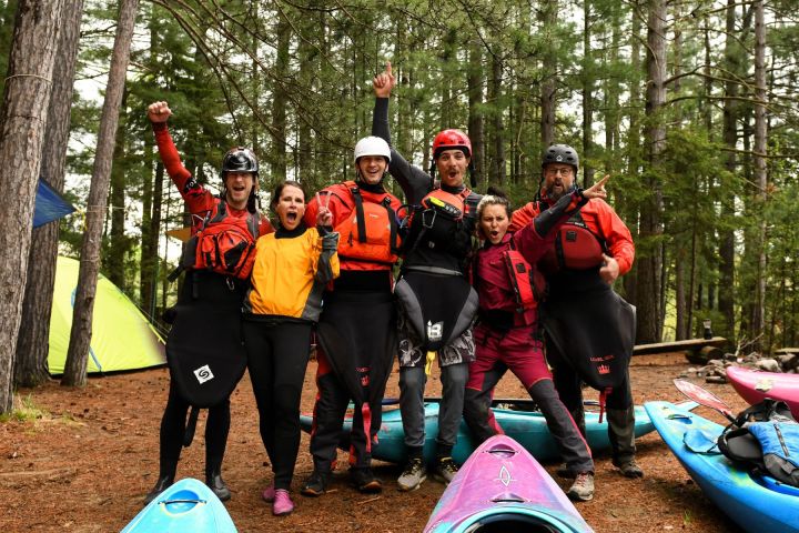 Group of six excited kayakers in gear, posing together in a forest campsite with kayaks.