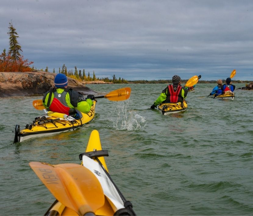 Four people kayaking on a lake surrounded by trees under a cloudy sky.