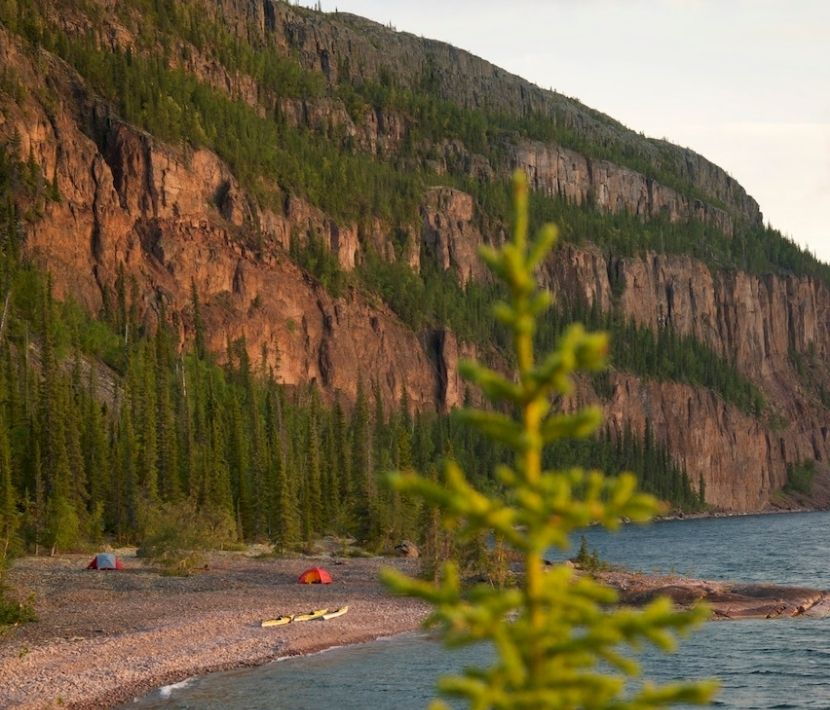 Rugged cliff with pine trees, tents, and kayaks by the lakeshore.