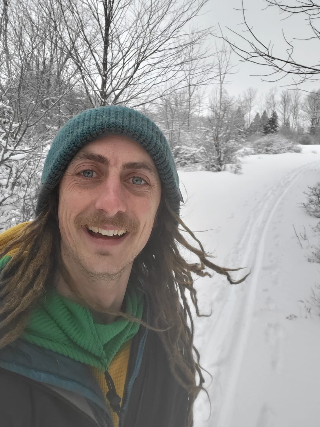 Person in a beanie smiles in a snowy landscape with bare trees.