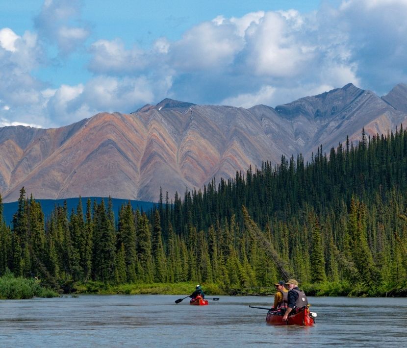 Canoes on a river with forest and mountain range in the background under a cloudy sky.