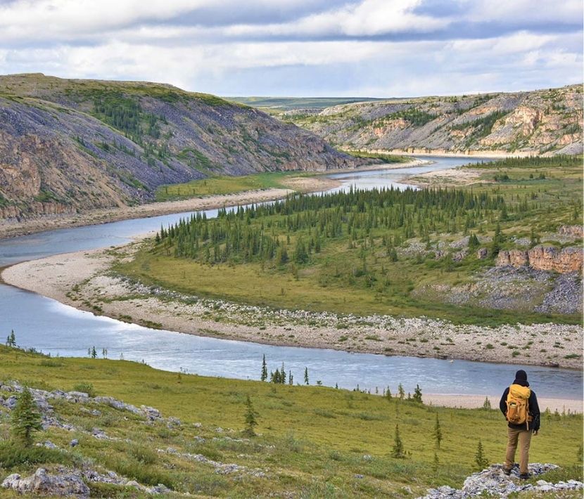 Person with backpack overlooks winding river and hilly landscape.