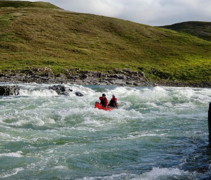 Two people in a red kayak navigating rough river rapids with grassy hills in the background.