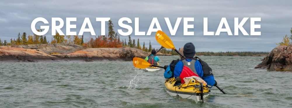Kayakers paddle on Great Slave Lake, surrounded by trees and rocky shores, under cloudy skies.