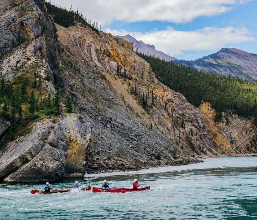 Four people kayaking on a river surrounded by rocky hills and trees under a partly cloudy sky.
