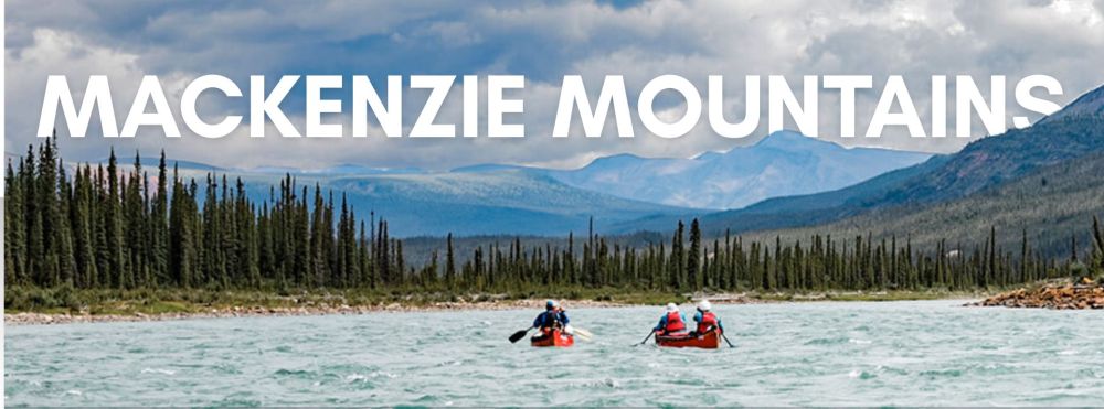 Two canoes on a river with evergreen trees and Mackenzie Mountains in the background.
