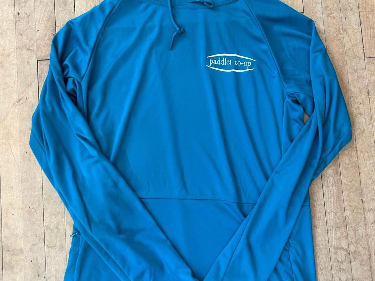 Blue hooded long-sleeve shirt with 'paddler co-op' logo on wood floor.