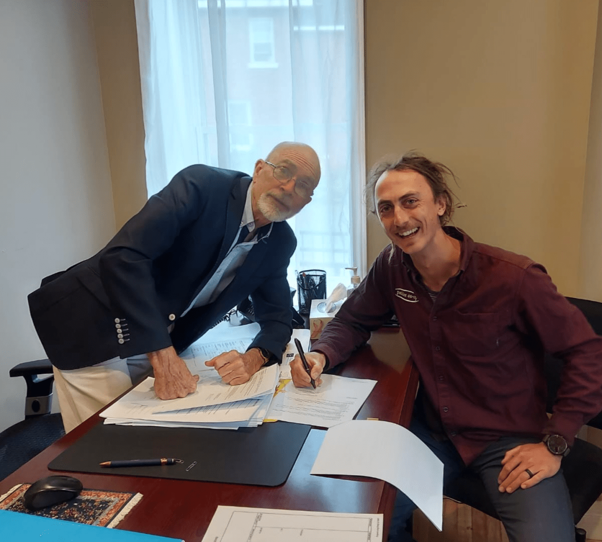 Two men smiling and signing papers at a desk in an office.